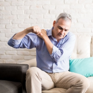 Older man in blue shirt and khakis sitting on couch holding his shoulder in pain