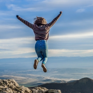 woman jumping with her arms up on top of a mountain summit